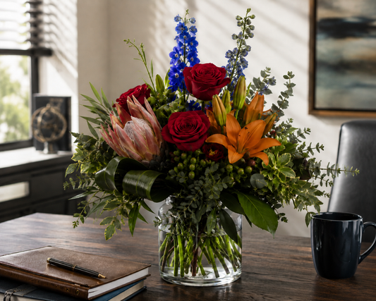 Masculine flower bouquet with red roses, orange lilies, protea, blue delphinium, greenery, and berries arranged in a glass vase on a wooden desk with a journal, pen, and black coffee mug